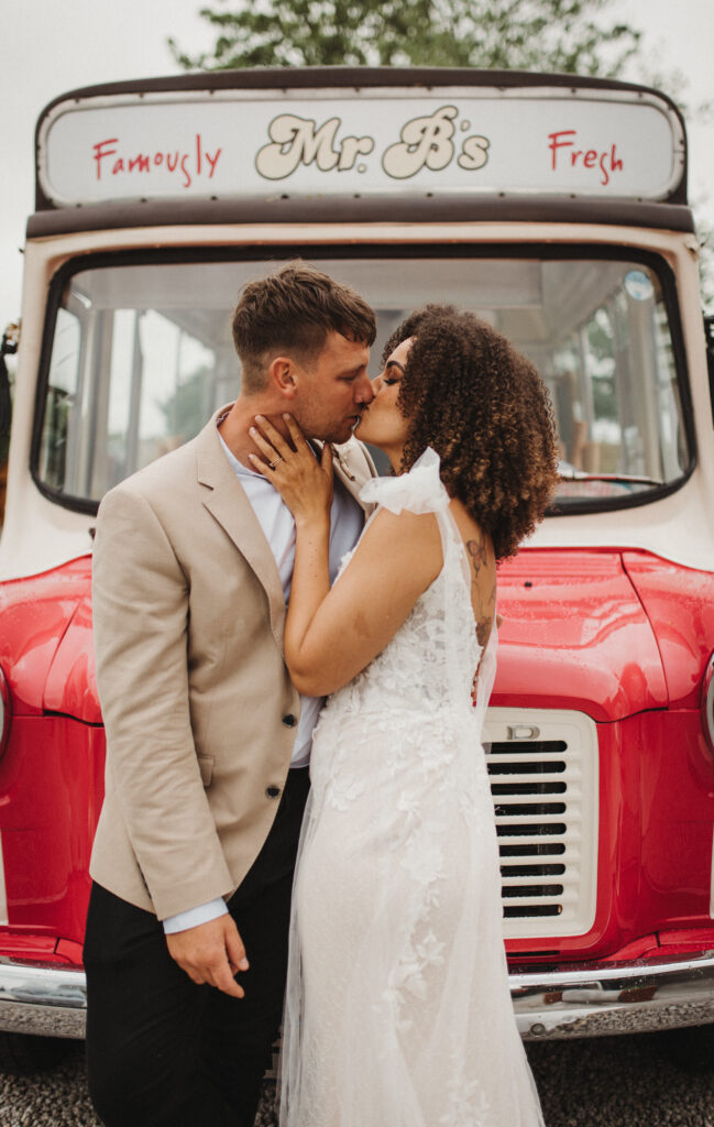 A curly-haired bride wearing her natural hair down kisses her groom in front of a vintage ice cream truck