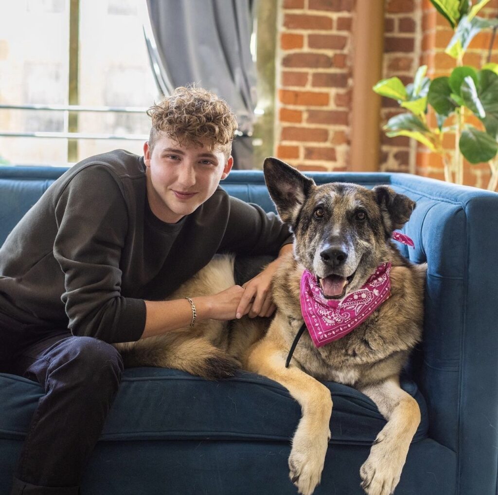 Matt Nelson, with short blond curly hair, smiles and cuddles with his black and tan German Shepherd Doug, wearing a pink bandana, on a blue velvet sofa. Doug has one ear standing up and one wonky ear, and also appears to be smiling.