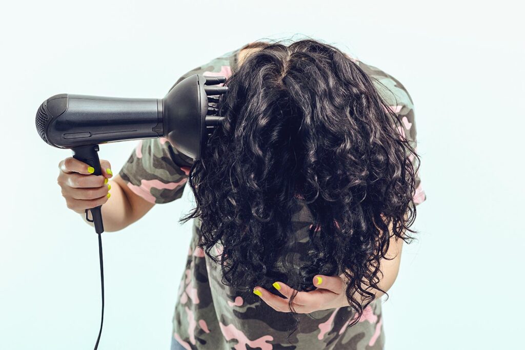 A woman with dark curly hair leans forward while diffusing her hair